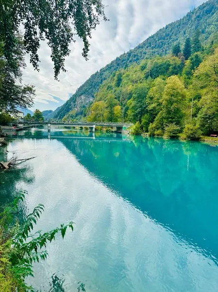 A breathtaking view of the Aare River flowing through Interlaken, with turquoise waters surrounded by lush greenery and majestic mountains in the background, under a clear blue sky.